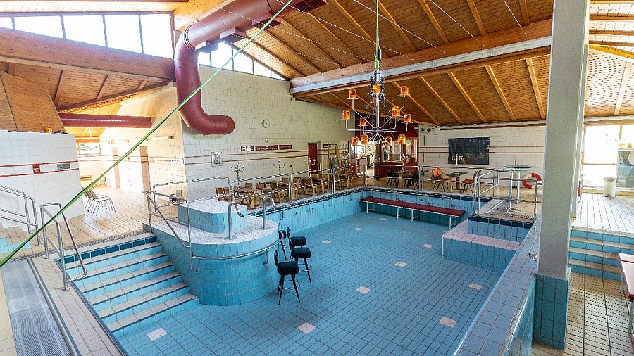 Das Bild zeigt das ehemalige Badehaus mit einem Pool in der Mitte und Tischen um den Pool herum. Der Raum wird jetzt für Kunst genutzt.
